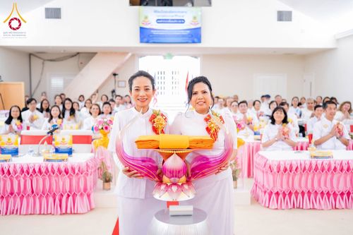 สุภาพสตรีสองท่านถือพานผ้าไตรจีวรสีเหลืองทองในพิธีทางศาสนาพุทธ โดยมีผู้เข้าร่วมพิธีนั่งพนมมืออยู่เบื้องหลัง
