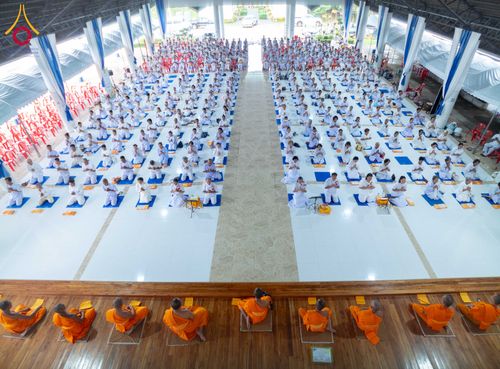 พระภิกษุและผู้เข้าร่วมโครงการอบรมปฏิบัติธรรมในชุดขาวนั่งเรียงแถวอย่างเป็นระเบียบภายในอาคารโถงกว้าง