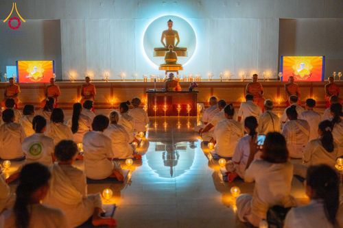 กลุ่มคนชุดขาวนั่งสมาธิในห้องโถงที่มีพระพุทธรูปและแสงเทียนประดับ