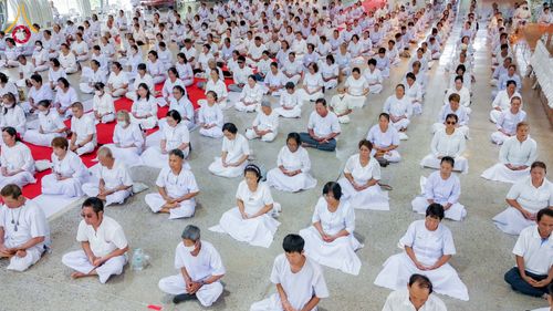 กลุ่มคนจำนวนมากสวมชุดสีขาวนั่งสมาธิเรียงแถวกันอย่างเป็นระเบียบภายในอาคาร