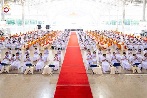 พุทธศาสนิกชนและพระภิกษุสงฆ์นั่งร่วมพิธีทางศาสนาภายในอาคารขนาดใหญ่ที่มีทางเดินปูพรมแดงตรงกลาง