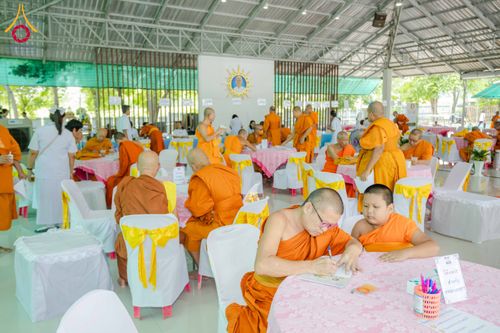 พระภิกษุและผู้เข้าร่วมงานกำลังนั่งทำกิจกรรมสัมมนาภายในอาคารที่มีการจัดโต๊ะเก้าอี้คลุมผ้าสีชมพูและสีขาว