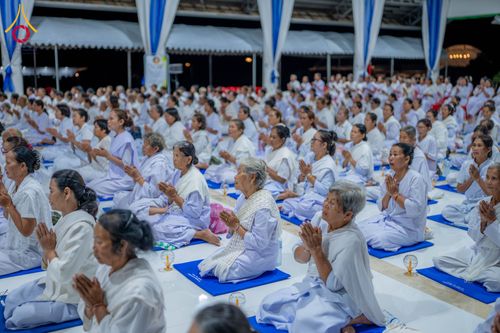 กลุ่มคนสวมชุดขาวนั่งพนมมือปฏิบัติธรรมบนเบาะรองนั่งสีน้ำเงินภายในอาคาร