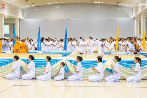 กลุ่มคนในชุดขาวนั่งพนมมือเรียงแถวหน้าโต๊ะวางพระของขวัญในพิธีทางศาสนา