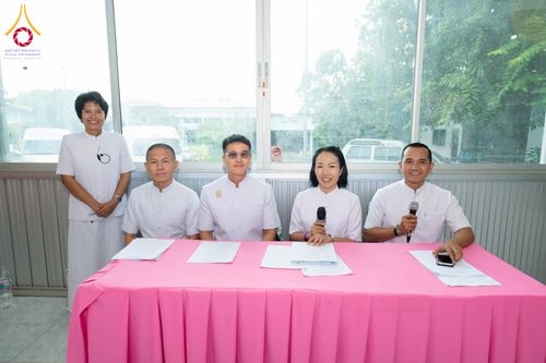 กลุ่มบุคคลชายหญิงสวมชุดขาวนั่งเรียงกันที่โต๊ะคลุมผ้าสีชมพูเพื่อเตรียมงาน
