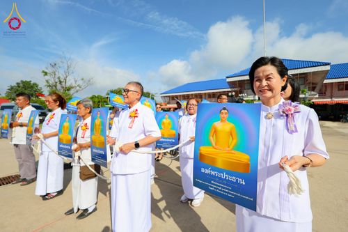กลุ่มคนสวมชุดขาวถือป้ายภาพพระประธานสีทองในขบวนพิธีกลางแจ้ง