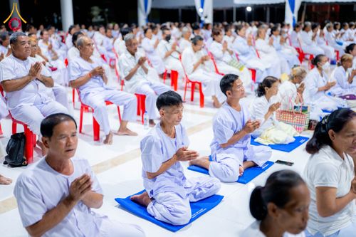 กลุ่มคนสวมชุดสีขาวนั่งพนมมือปฏิบัติธรรมบนเบาะรองนั่งภายในอาคาร