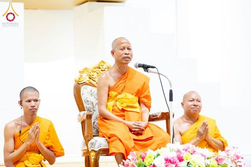 พระสงฆ์สามรูปในจีวรสีส้มกำลังประกอบพิธีทางศาสนา โดยรูปกลางนั่งบนเก้าอี้ไม้แกะสลักและกำลังพูดผ่านไมโครโฟน