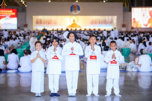 เด็กชายและเด็กหญิง 5 คนสวมชุดสีขาวพนมมือไหว้ในอาคารที่มีผู้คนร่วมพิธีตักบาตรอยู่ด้านหลัง