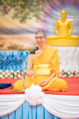 พระภิกษุสงฆ์กำลังนั่งเทศนาธรรมบนเวทีที่มีพระพุทธรูปเป็นฉากหลัง