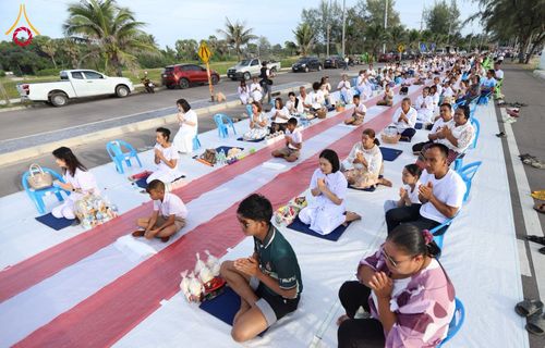 กลุ่มประชาชนสวมชุดสีขาวนั่งพนมมือบนผืนผ้าสีขาวแดงริมถนนติดชายหาดเพื่อรอตักบาตร