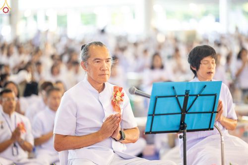 ชายวัยกลางคนสวมชุดขาวกำลังพนมมือสวดมนต์นำกลุ่มพุทธศาสนิกชนที่นั่งอยู่ด้านหลังในพิธีกรรมทางศาสนา