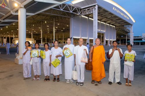 กลุ่มคนสวมชุดขาวและพระภิกษุสงฆ์ถือถุงอาหารแห้งยืนเรียงกันหน้าอาคารสภาธรรมกายสากล