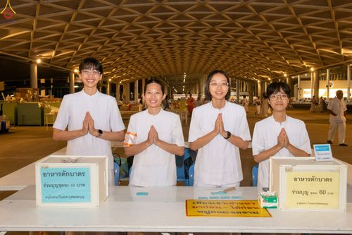 อาสาสมัครหญิงสี่คนสวมชุดสีขาวพนมมือยืนประจำจุดบริการอาหารตักบาตรภายในอาคารขนาดใหญ่