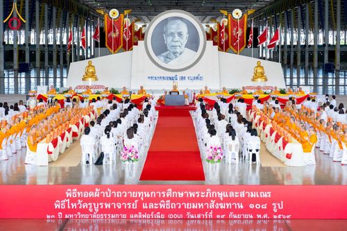 พุทธศาสนิกชนและพระภิกษุสงฆ์จำนวนมากเข้าร่วมพิธีทางศาสนาภายในสภาธรรมกายสากล โดยมีฉากหลังเป็นภาพพระมงคลเทพมุนีและธงประดับตกแต่งอย่างสวยงาม