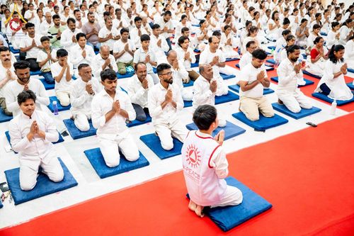 กลุ่มคนสวมชุดสีขาวนั่งพนมมือปฏิบัติธรรมบนเบาะรองนั่งในอาคาร โดยมีเจ้าหน้าที่สวมเสื้อกั๊กสีขาวนั่งหันหลังอยู่ด้านหน้า