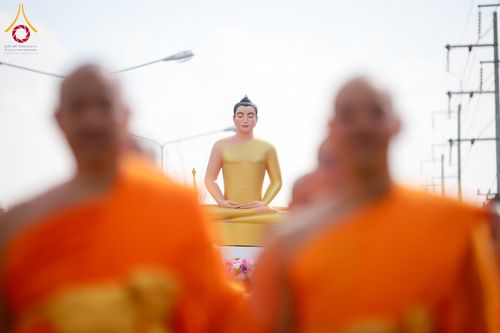 พระพุทธรูปสีทองปางสมาธิโดยมีพระภิกษุสงฆ์ในจีวรสีส้มเดินผ่านอยู่ด้านหน้าในลักษณะภาพเบลอ