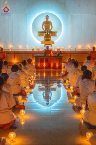กลุ่มผู้เข้าร่วมโครงการสวมชุดขาวนั่งปฏิบัติธรรมภายในอาคารที่มีแสงเทียนวางเรียงรายบนพื้นและมีพระภิกษุนั่งเป็นประธานด้านหน้า