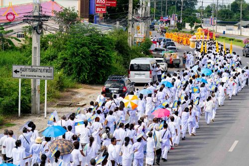 กลุ่มคนสวมชุดขาวจำนวนมากกำลังเดินขบวนไปตามถนนในพิธีทางศาสนา โดยมีขบวนพระภิกษุสงฆ์และรถบรรทุกองค์พระประธานนำหน้า
