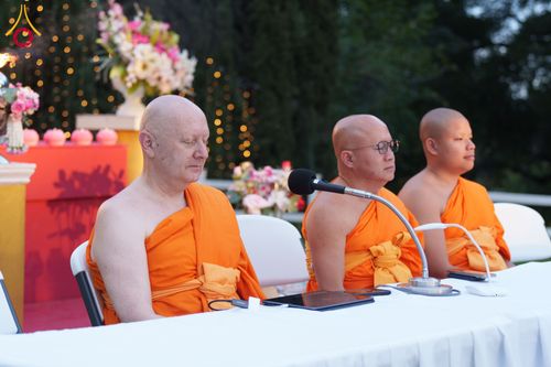 พระภิกษุสงฆ์สามรูปนั่งสมาธิอยู่หลังโต๊ะที่มีไมโครโฟน ในบรรยากาศงานพิธีกลางแจ้ง