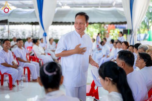 ชายวัยกลางคนสวมชุดสีขาวกำลังยืนยิ้มและพูดคุยกับกลุ่มผู้เข้าอบรมที่นั่งอยู่ภายในเต็นท์ปฏิบัติธรรม