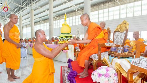 พระภิกษุสงฆ์ในจีวรสีส้มกำลังประกอบพิธีทางศาสนาและถือพานพุ่มเกศดอกบัวตูมสีเหลืองทองภายในอาคาร