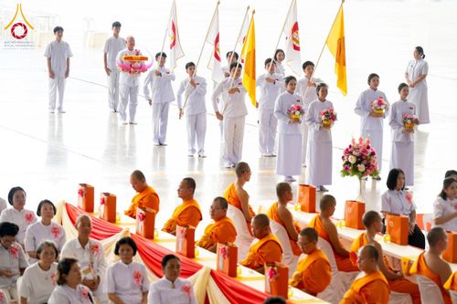ขบวนผู้ถือธงและพานดอกไม้เดินนำหน้าพระภิกษุสงฆ์และพุทธศาสนิกชนชุดขาวในพิธีทางศาสนา