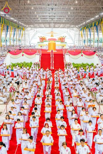 พุทธศาสนิกชนชุดขาวจำนวนมากยืนถือพานพุ่มในพิธีทอดผ้าป่าภายในอาคารขนาดใหญ่ที่มีพรมแดงและเวทีประดิษฐานพระพุทธรูป