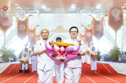 ชายสองคนสวมชุดขาวถือพานพุ่มผ้าไตรจีวรเดินนำขบวนในพิธีทางศาสนา