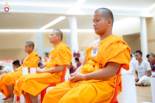 พระภิกษุสงฆ์หลายรูปนั่งสมาธิหลับตาในจีวรสีเหลืองทองภายในห้องโถง