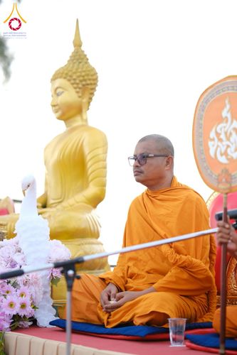 พระภิกษุสงฆ์สวมจีวรสีส้มและแว่นตานั่งอยู่หน้าพระพุทธรูปสีทองในพิธีทางศาสนา