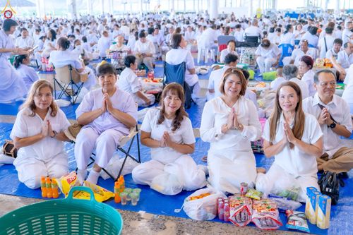 กลุ่มคนสวมชุดขาวนั่งพนมมือบนพื้นในอาคารกว้างขวางเพื่อร่วมพิธีตักบาตร