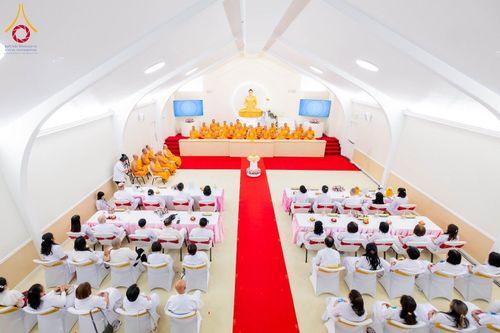 พระภิกษุสงฆ์และสาธุชนชุดขาวร่วมพิธีทางศาสนาภายในอาคารปฏิบัติธรรมที่มีพระประธานเป็นศูนย์กลาง