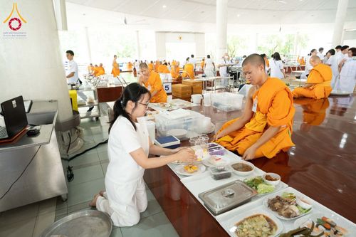 หญิงชุดขาวกำลังประเคนอาหารแด่พระภิกษุสงฆ์ภายในหอฉันที่มีผู้คนร่วมงานจำนวนมาก