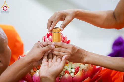 มือของพระภิกษุและฆราวาสกำลังประคองตลับสีทองเหนือพานพุ่มดอกบัว