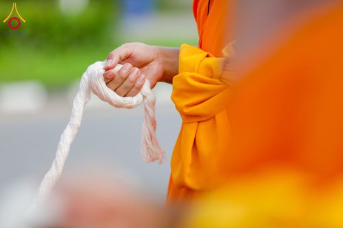 มือพระภิกษุสวมจีวรสีส้มกำลังถือสายสิญจน์สีขาวในพิธีกรรม
