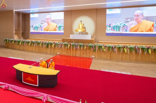 พระภิกษุสงฆ์บนเวทีในห้องประชุมที่มีจอภาพขนาดใหญ่และพระพุทธรูป