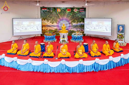 พระภิกษุสงฆ์นั่งสมาธิบนเวทีในพิธีทางศาสนาที่มีพระพุทธรูปและจอโปรเจกเตอร์เป็นฉากหลัง