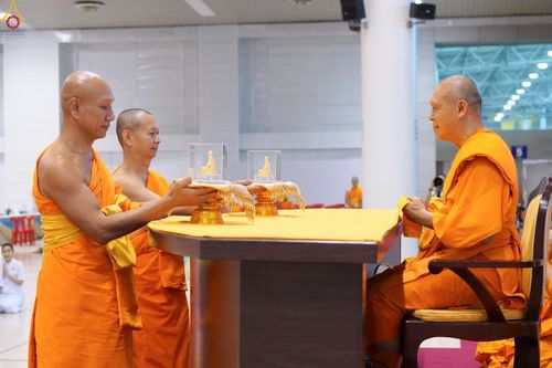 พระภิกษุสงฆ์ในจีวรสีส้มกำลังประกอบพิธีมอบพระของขวัญที่วางอยู่บนพานทองภายในห้องโถง