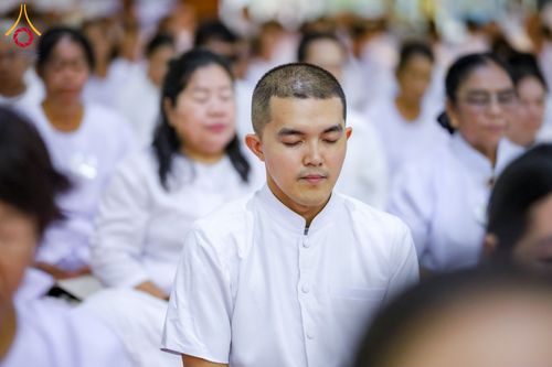 ชายหนุ่มและกลุ่มผู้เข้าร่วมโครงการปฏิบัติธรรมสวมชุดสีขาวกำลังนั่งหลับตาทำสมาธิ