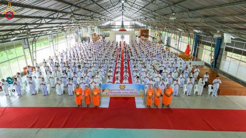 กลุ่มผู้เข้าร่วมโครงการอบรมในชุดสีขาวนั่งเรียงแถวภายในอาคารโดยมีพระภิกษุสงฆ์ยืนอยู่ด้านหน้า