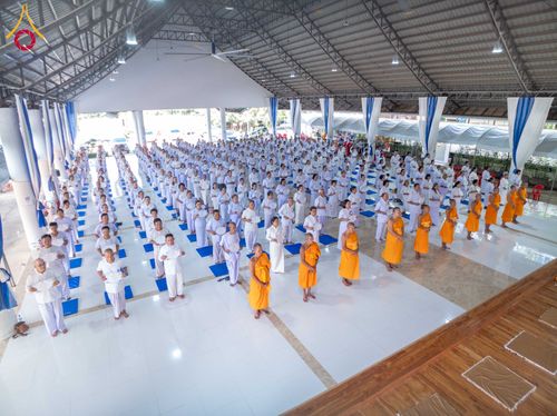 กลุ่มผู้เข้าร่วมโครงการอบรมสวมชุดสีขาวและพระภิกษุสวมจีวรสีเหลืองยืนเรียงแถวทำกิจกรรมภายในอาคาร