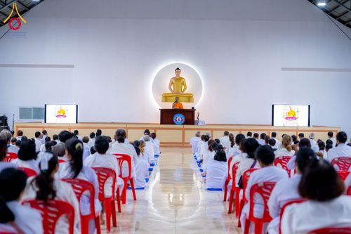 กลุ่มผู้เข้าร่วมอบรมสวมชุดขาวนั่งฟังธรรมจากพระภิกษุบนเวทีภายในอาคาร