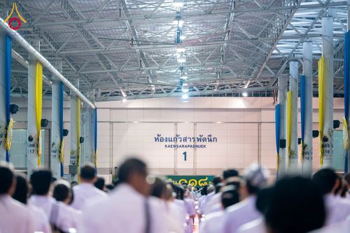 กลุ่มคนสวมชุดสีขาวนั่งรวมตัวกันภายในอาคารขนาดใหญ่ที่มีป้ายชื่อห้องแก้วสารพัดนึก 1 ติดอยู่ที่ผนังด้านหน้า
