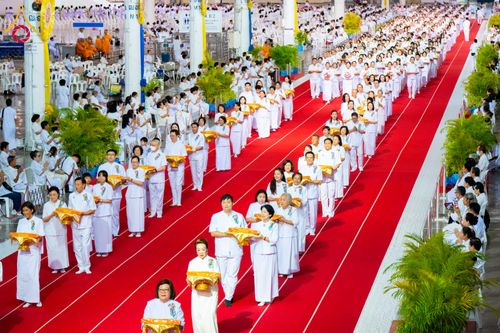 กลุ่มพุทธศาสนิกชนสวมชุดขาวถือพานพุ่มเดินแถวบนพรมแดงในอาคารขนาดใหญ่