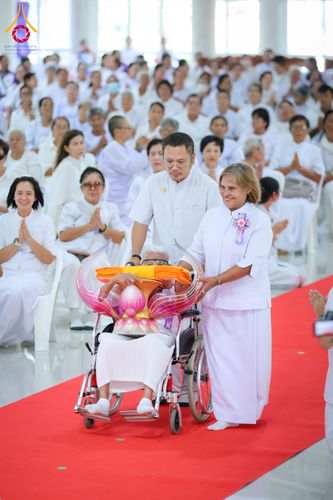 กลุ่มคนในชุดขาวกำลังเข็นรถเข็นที่มีพานพุ่มวางผ้าไตรสีเหลืองทองบนพรมสีแดงภายในอาคาร