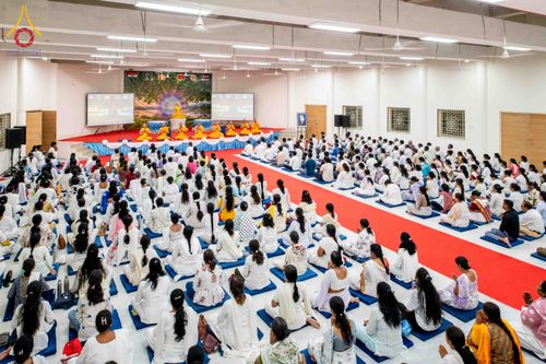 ผู้คนจำนวนมากสวมชุดสีขาวนั่งเรียงแถวปฏิบัติธรรมภายในอาคารที่มีพระภิกษุสงฆ์อยู่บนเวทีด้านหน้า