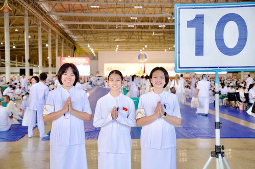 หญิงสาวสามคนสวมชุดสีขาวพนมมือยิ้มแย้มในโถงกว้างที่มีผู้คนร่วมกิจกรรมทางศาสนาและมีป้ายหมายเลข 10