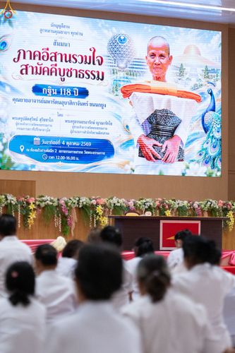 ผู้เข้าร่วมงานสัมมนาในชุดสีขาวนั่งหันหลังให้กล้อง มองไปยังจอภาพขนาดใหญ่ที่แสดงรายละเอียดงานกฐิน 118 ปี คุณยายอาจารย์มหารัตนอุบาสิกาจันทร์ ขนนกยูง