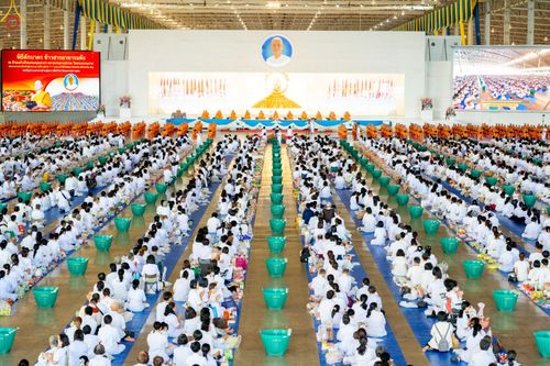พุทธศาสนิกชนสวมชุดขาวนั่งเรียงแถวร่วมพิธีตักบาตรภายในอาคารขนาดใหญ่ที่มีพระภิกษุสงฆ์นั่งอยู่ด้านหน้า